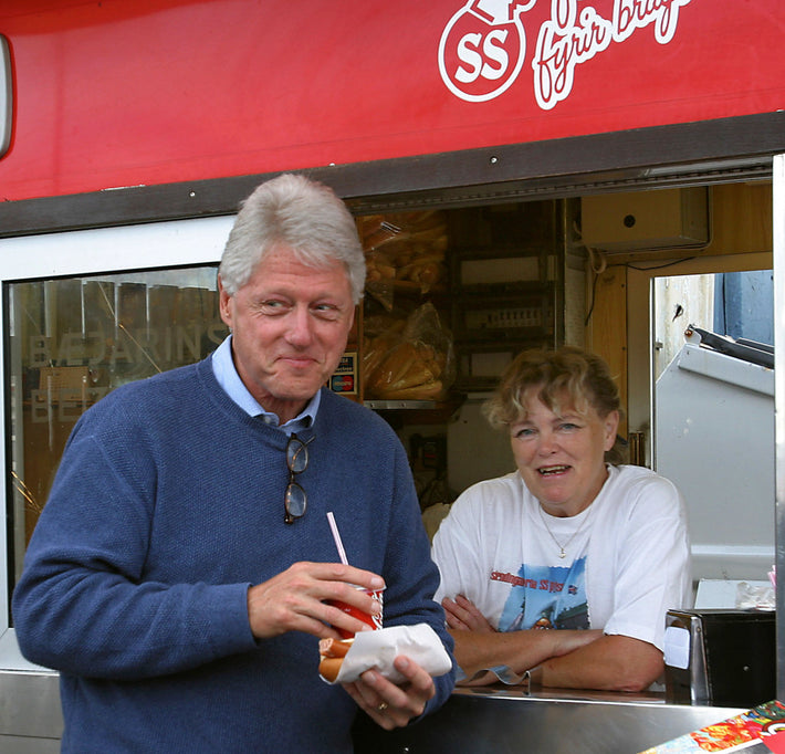 A man holding a hot dog and drink stands in front of a food stall, smiling, while a woman behind the counter also smiles. The stall has a red awning with text in Icelandic.