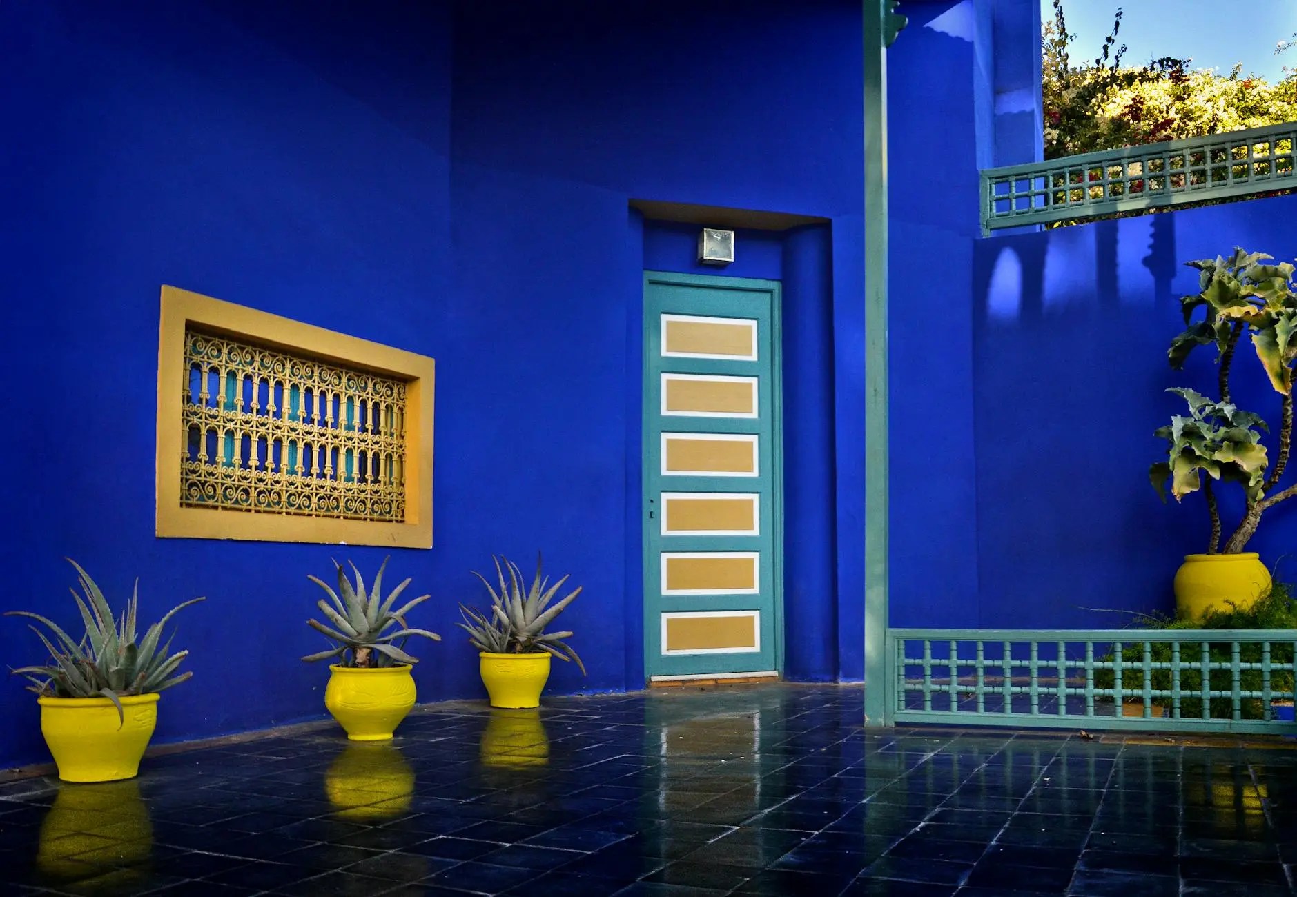 A vibrant blue wall with a decorative yellow window frame and a green door. Several yellow pots with agave plants are placed in front, reflecting on the shiny black floor.