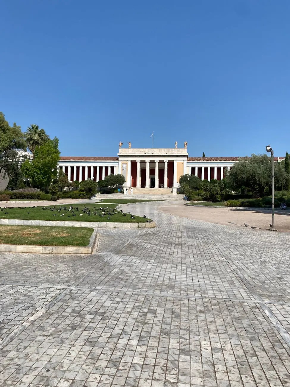 View of a classical building surrounded by greenery under a clear blue sky, featuring tall columns and a spacious plaza with scattered birds.