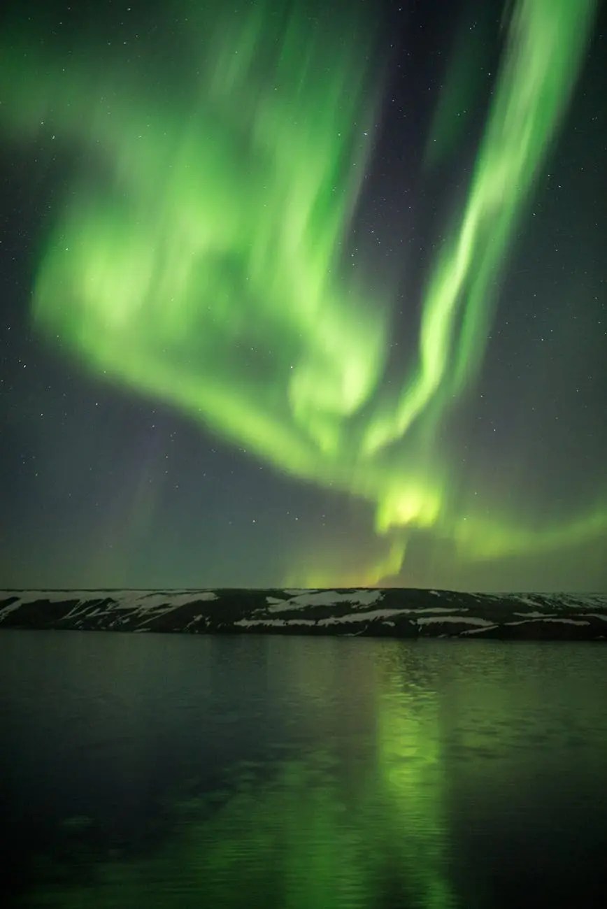 A vibrant display of the Northern Lights illuminating the night sky over a dark landscape with a calm reflection on water.