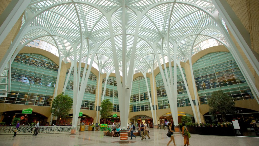 Interior view of a modern shopping complex with a high, white lattice ceiling and large glass windows, showcasing plants and visitors walking through the space.