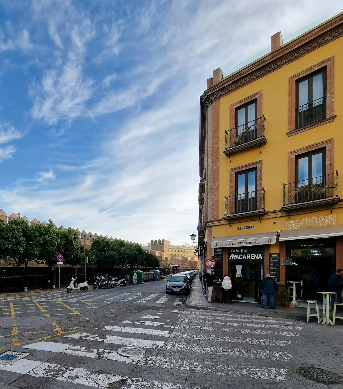 A street view in Seville featuring a vibrant yellow building with balconies, a cafe-bar named 'Macarena', and orange trees lining the street under a clear blue sky.