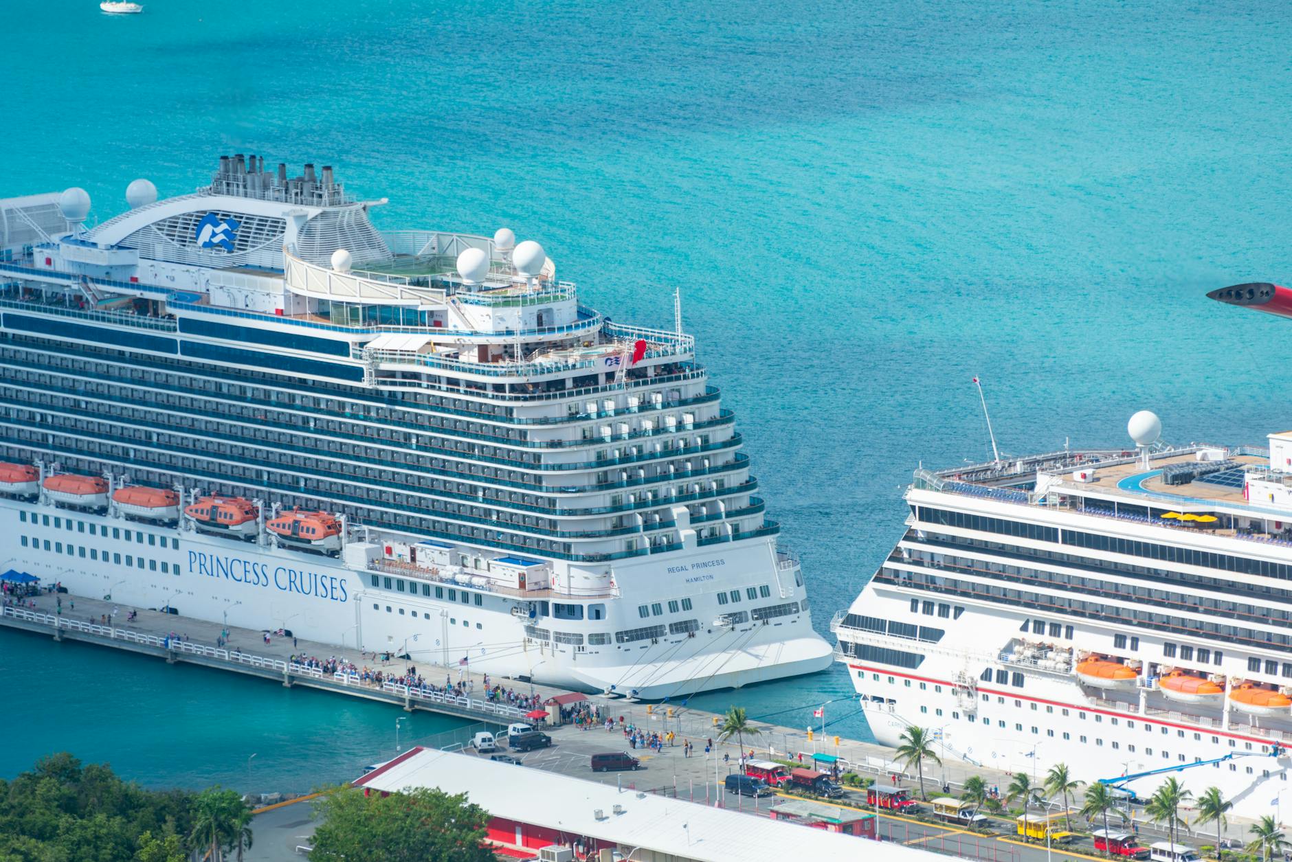 Two large cruise ships docked at a port with turquoise water in the background.