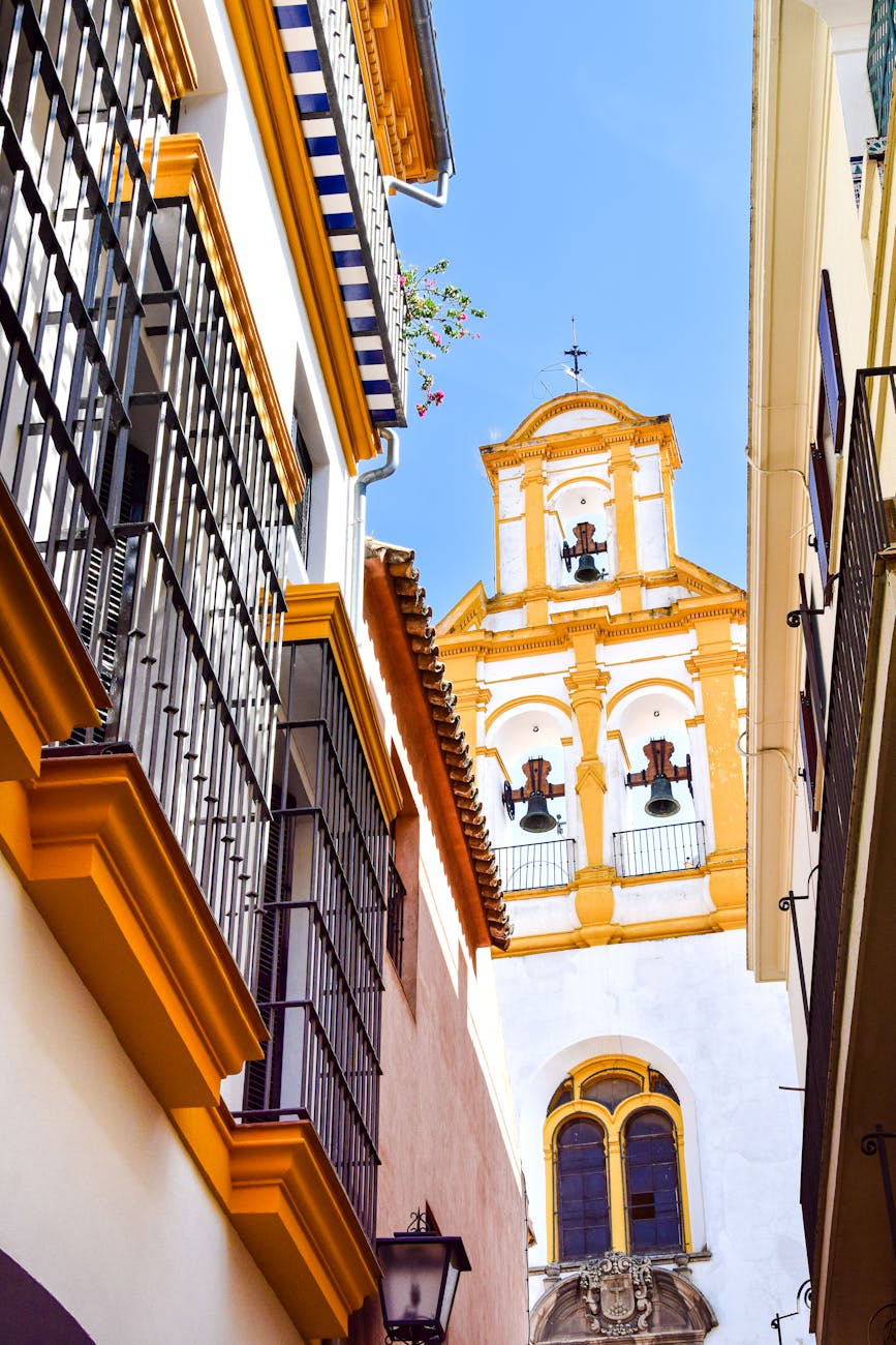 A narrow street in Seville, Spain, showcasing traditional white and yellow architecture, with a bell tower visible in the background under a clear blue sky.