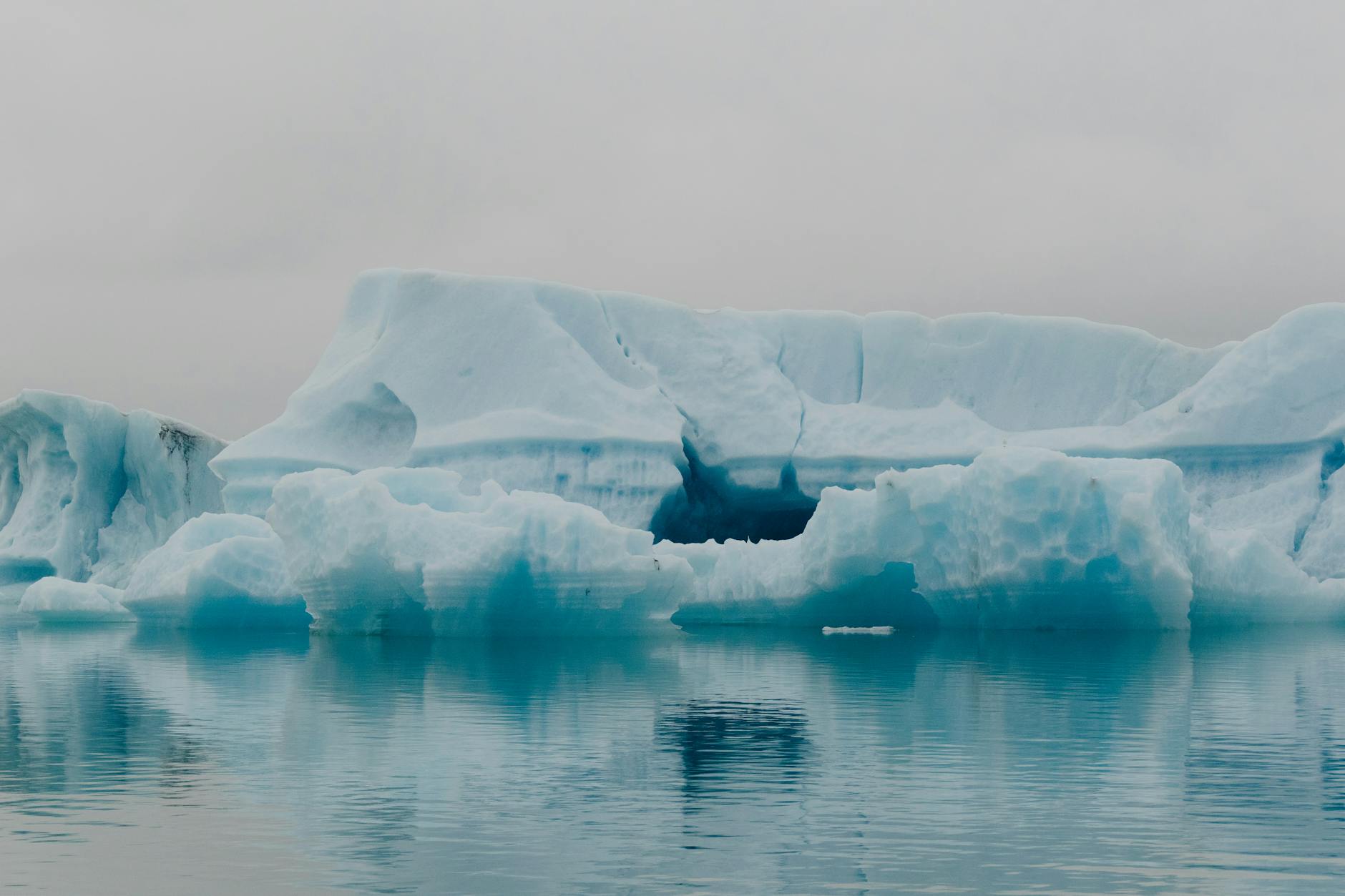A large iceberg floats in calm water, reflecting shades of blue and white, under a gray sky.