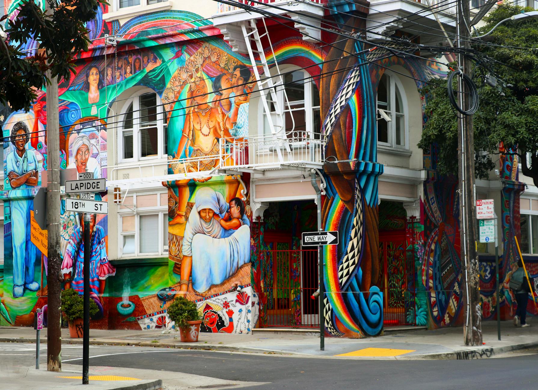 A vibrant mural depicting cultural scenes and figures on a building in San Francisco's Mission District, featuring colorful artwork and intricate designs.