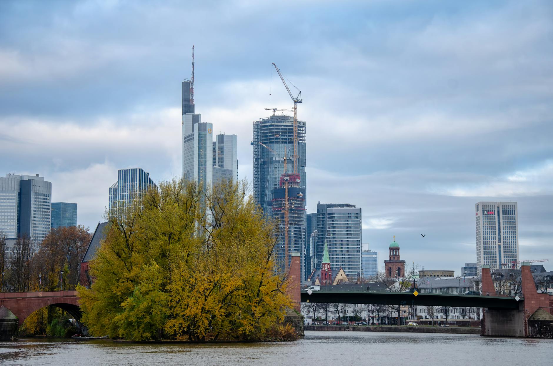 A skyline view of Frankfurt am Main featuring modern skyscrapers, a crane, and a green park along the riverbank, with a cloudy sky overhead.