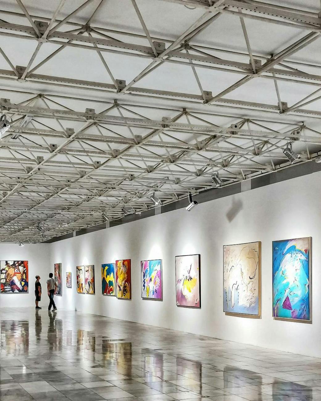 Interior view of an art gallery showcasing colorful abstract paintings on white walls, with two visitors walking along a polished floor.