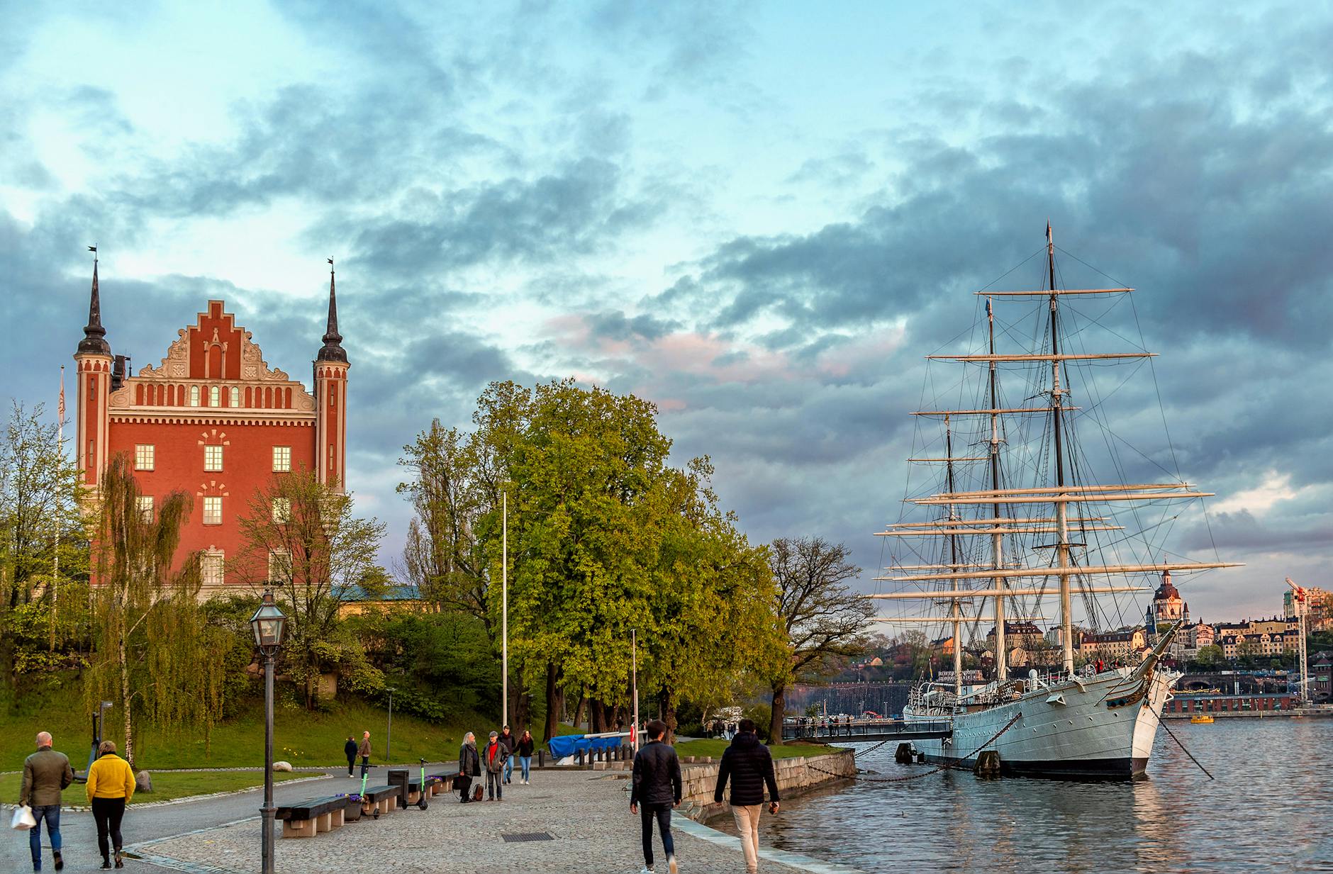 A scenic view of Stockholm's waterfront featuring a vintage ship docked beside a colorful historic building, with people walking along the promenade and lush trees along the water's edge.