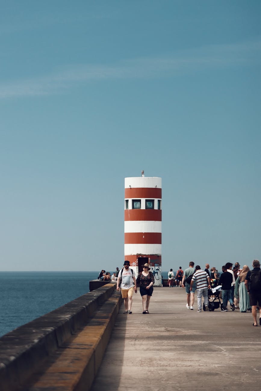 A busy seaside pier with people walking and enjoying the view, featuring a striped lighthouse at the end and a clear blue sky in the background.