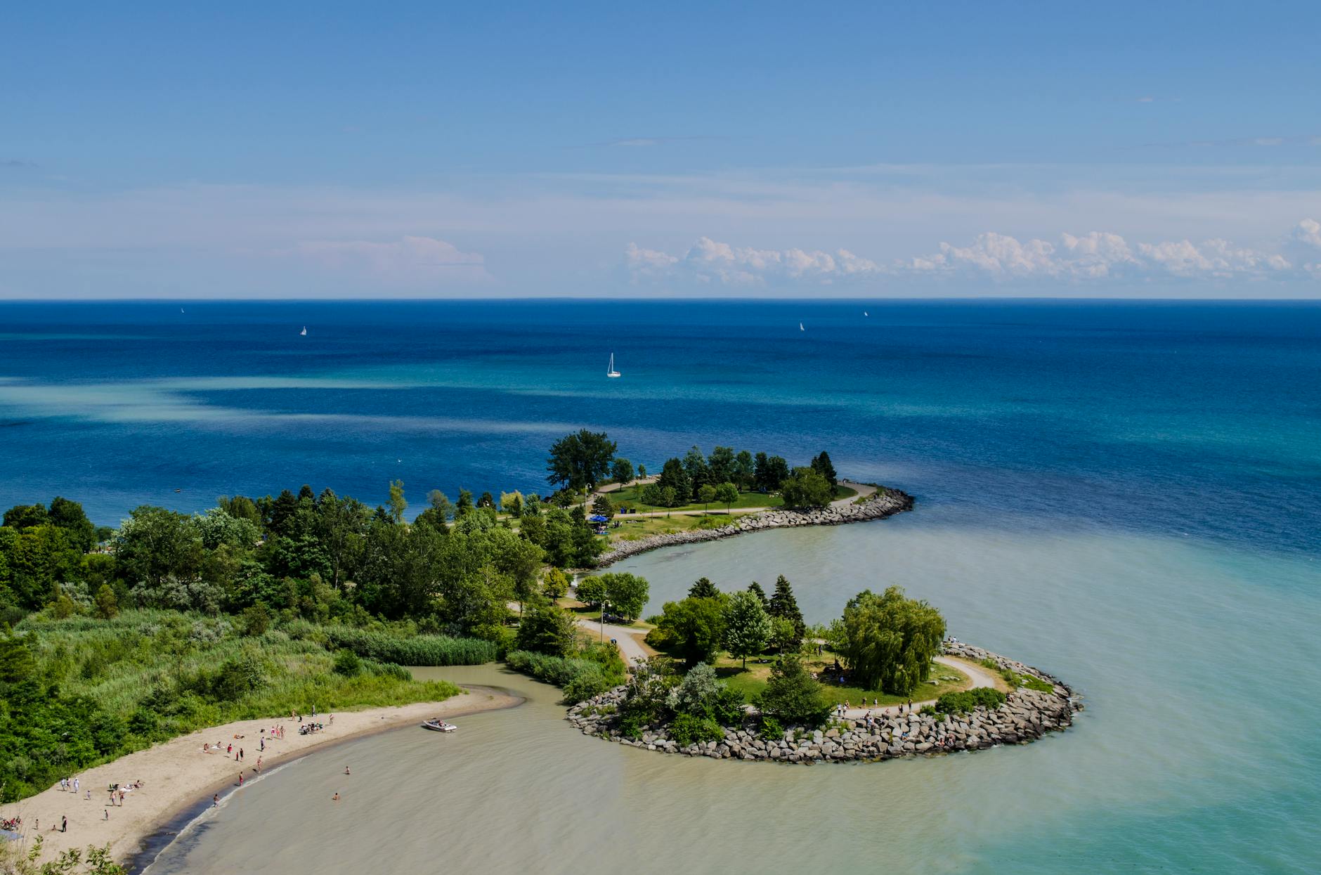 A serene view of a coastal area with a sandy beach, surrounded by lush greenery and azure waters, featuring small boats and sailboats in the distance.