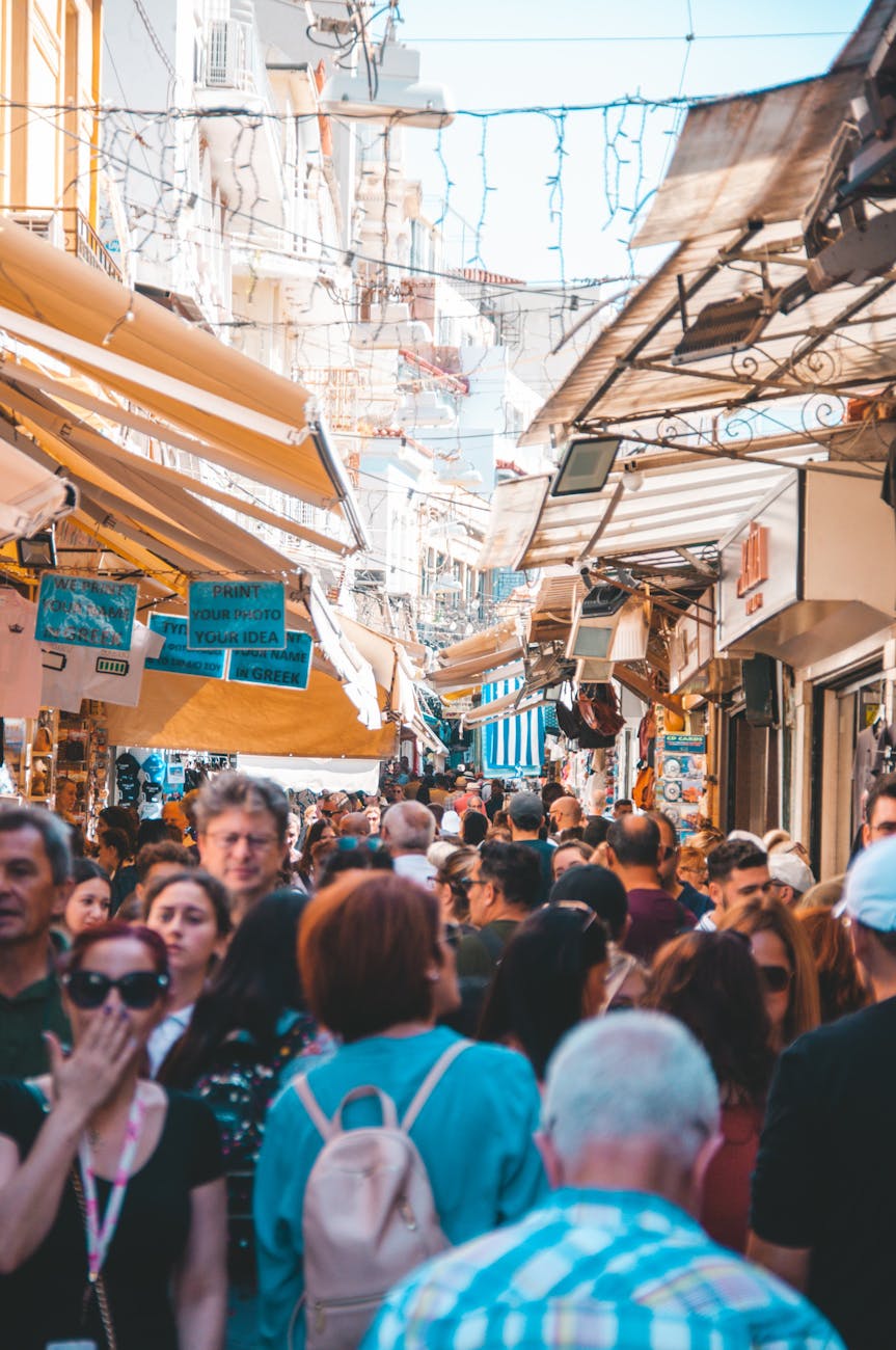 A bustling street market in Athens filled with people exploring shops and stalls, with colorful awnings overhead and various items displayed for sale.
