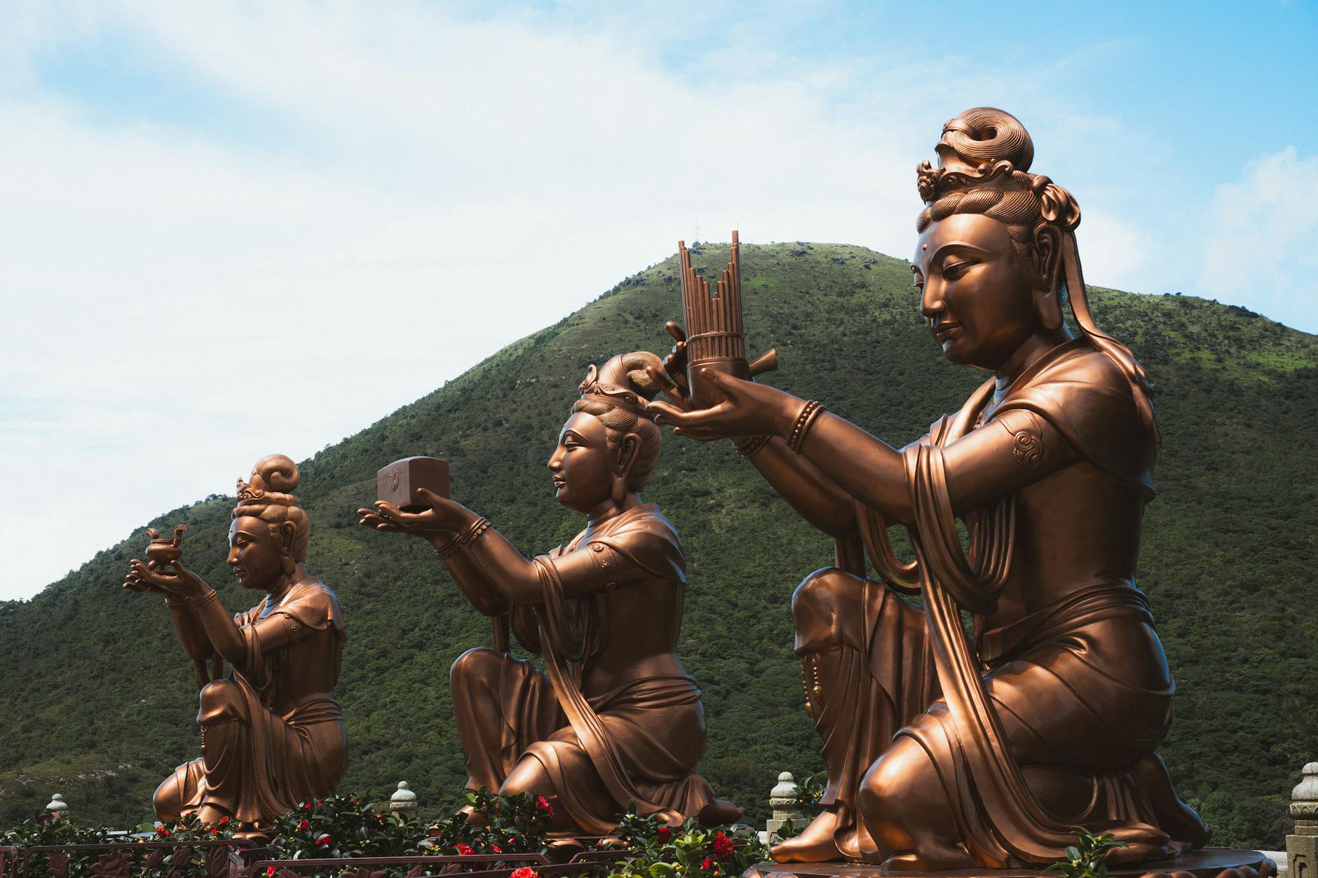 Three bronze statues of deities holding offerings, set against a backdrop of green mountains under a blue sky.