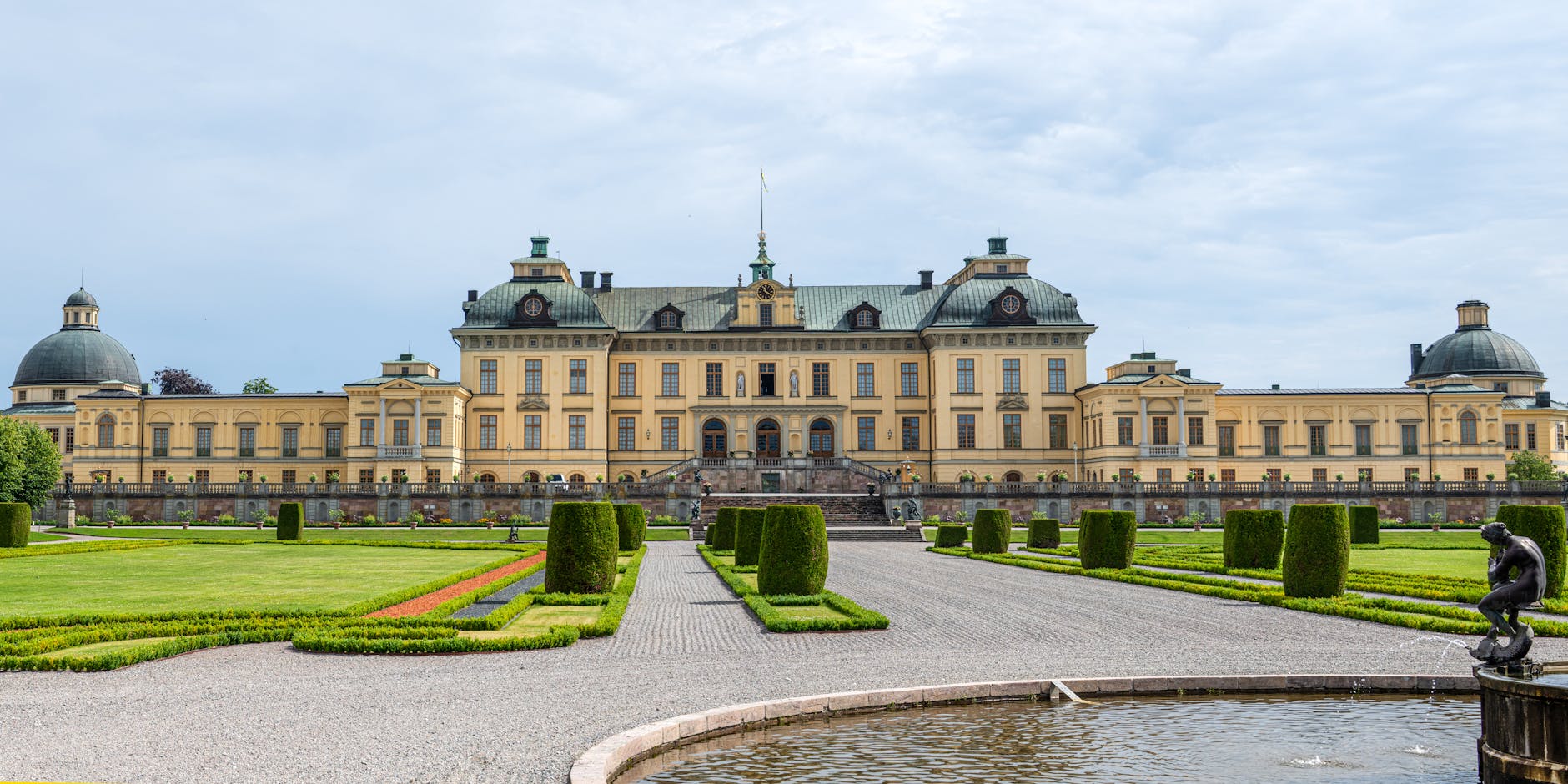 A grand historic palace with a manicured garden in the foreground, featuring neatly trimmed hedges and a statue by the pond, under a cloudy sky.