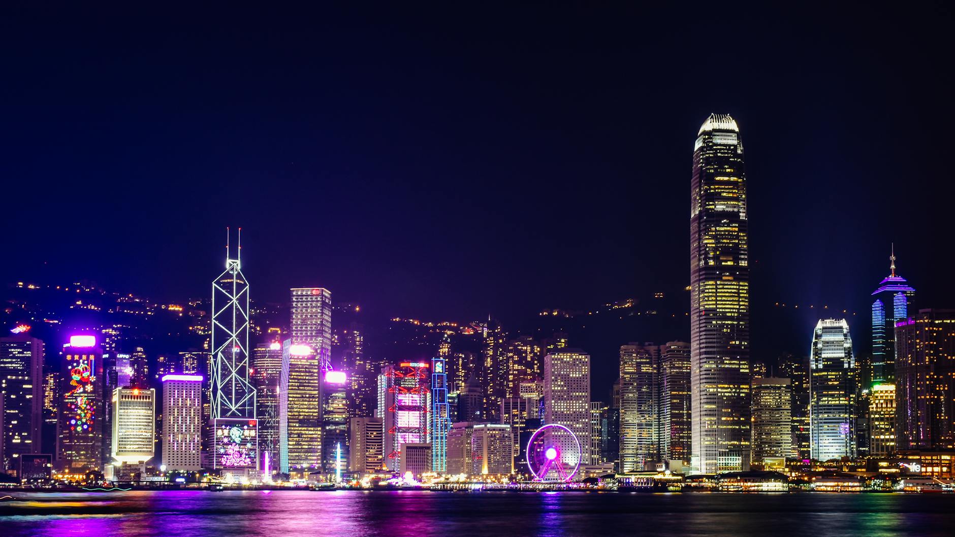 A panoramic view of Hong Kong's skyline at night, showcasing illuminated skyscrapers and the iconic Ferris wheel.
