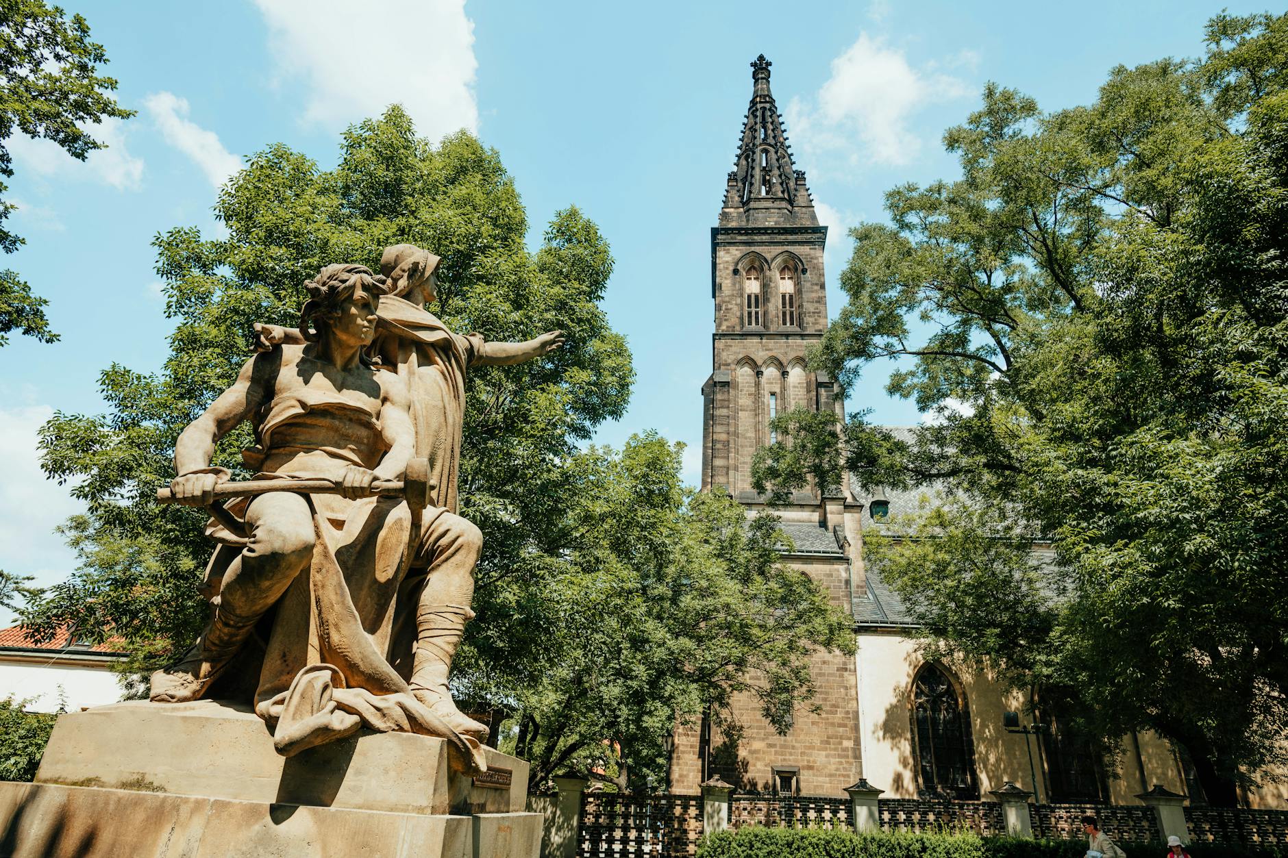 A close-up of a statue of two figures, one pointing forward, with a tall, ornate church tower in the background, surrounded by lush green trees against a blue sky.