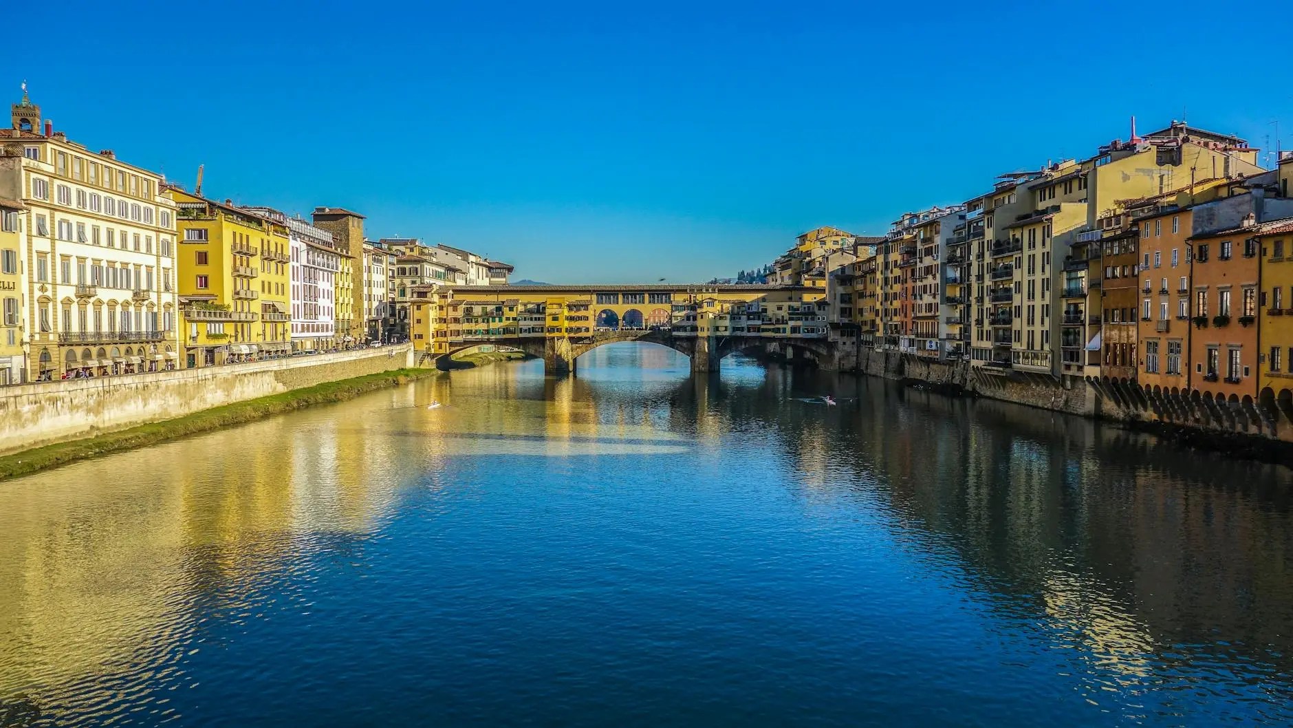 Scenic view of the Ponte Vecchio bridge over the Arno River in Florence, Italy, with colorful buildings lining the riverbanks under a clear blue sky.