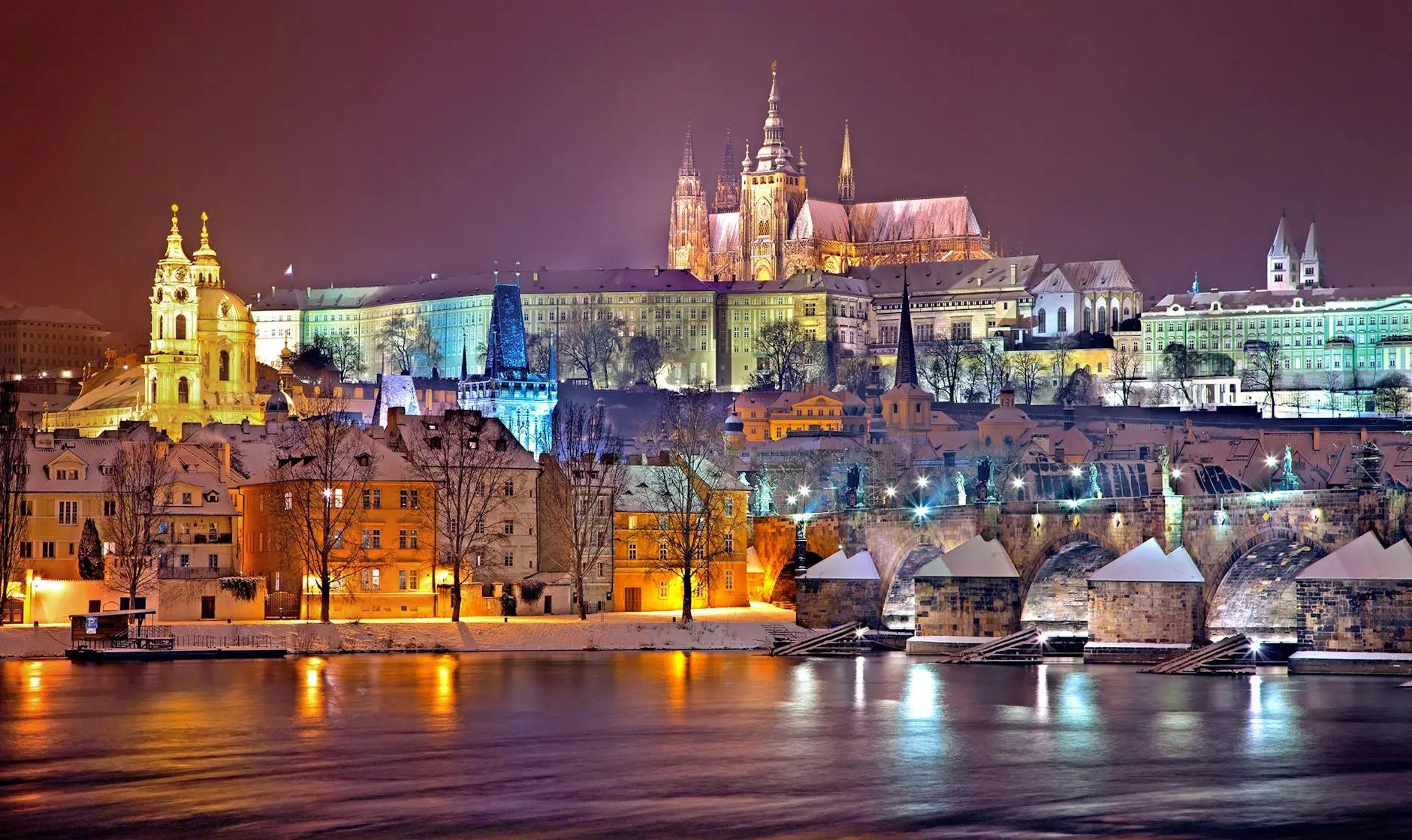 Night view of Prague Castle illuminated above the Vltava River, with historic buildings and the Charles Bridge in the foreground under a snowy sky.