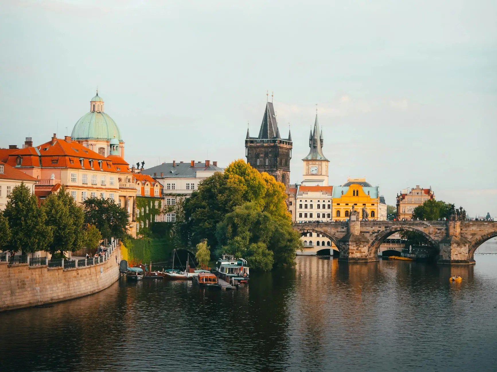 A scenic view of Prague, showcasing historic buildings with red rooftops along the Vltava River, featuring the iconic Charles Bridge in the foreground.
