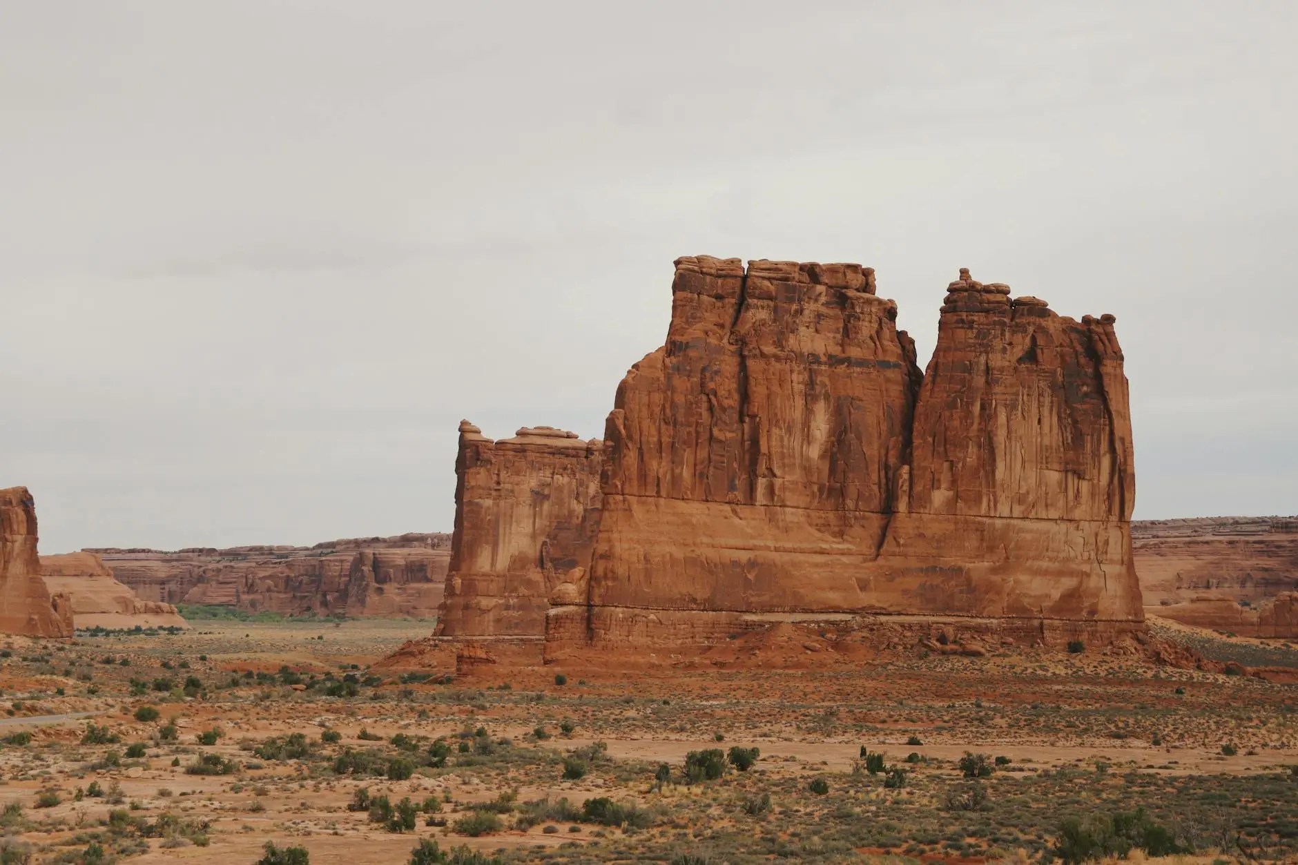 A scenic view of large red rock formations against a cloudy sky, surrounded by desert terrain and sparse vegetation.