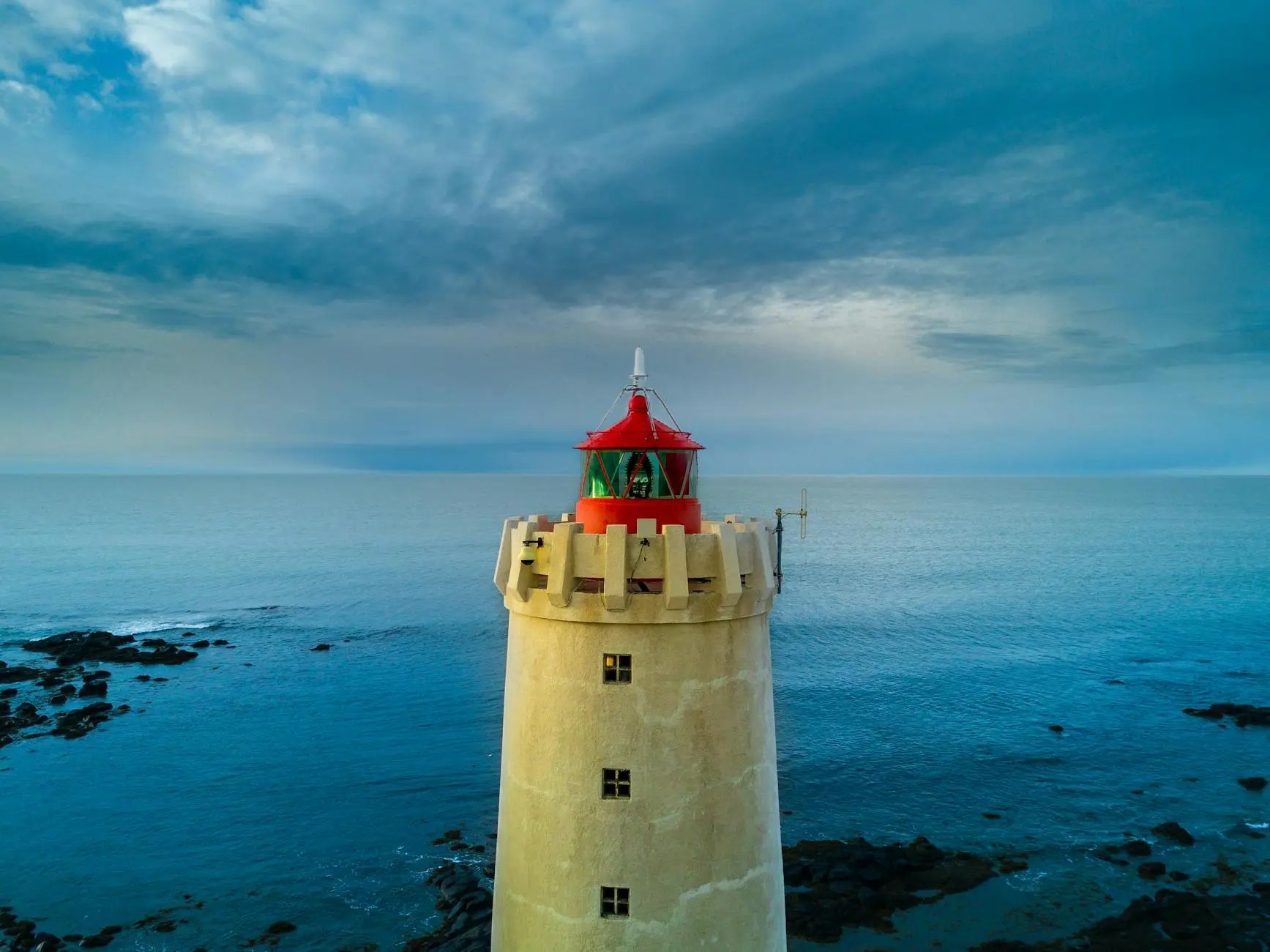 A close-up view of a lighthouse with a red top, standing against a backdrop of a serene ocean and dramatic cloudy sky.