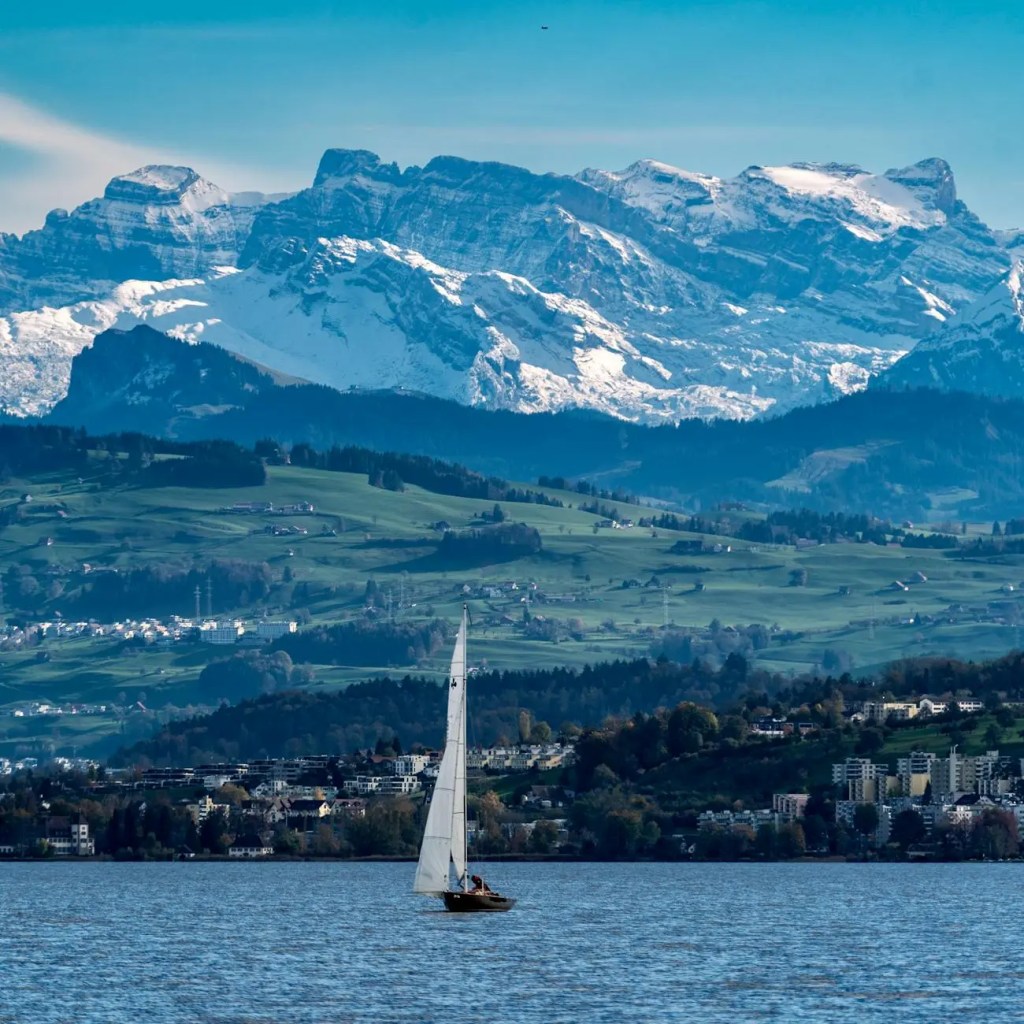 Sailboat gliding on Lake Zurich with snow-capped mountains in the background, showcasing a picturesque landscape.
