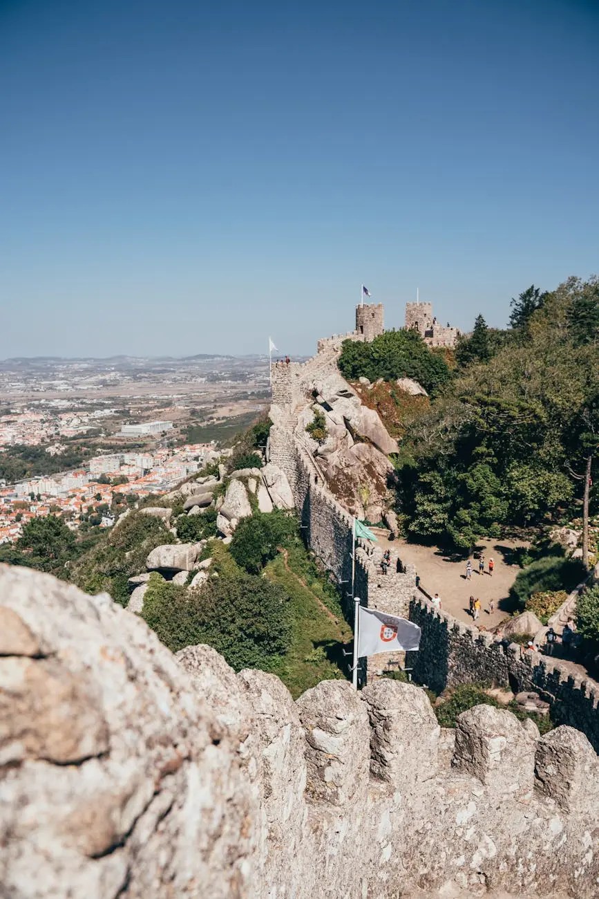 View from the Castle of São Jorge in Lisbon, showing ancient stone walls, flags, and a panoramic landscape of the city and surrounding hills under a clear blue sky.