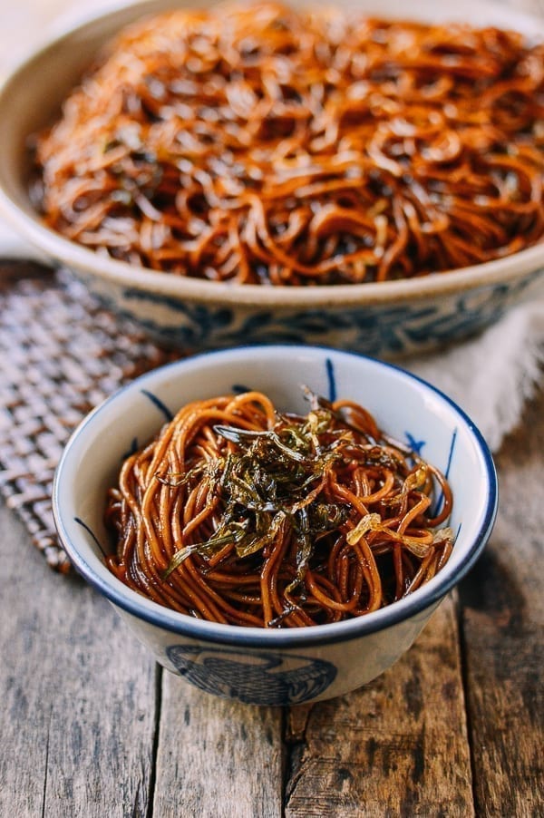 A bowl of dark, glossy noodles topped with finely chopped herbs, placed on a rustic wooden table, with a larger serving dish of the same noodles in the background.