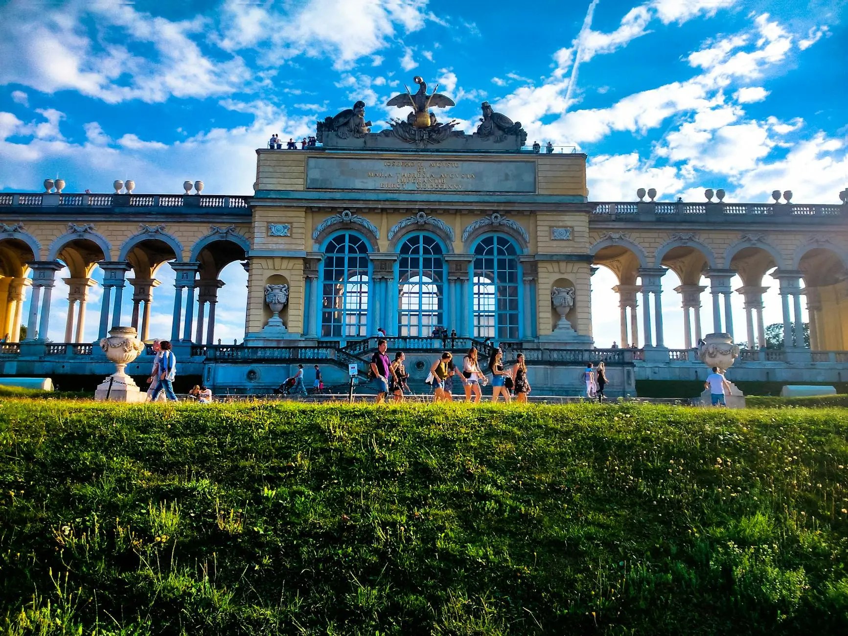View of the Gloriette at Schönbrunn Palace, with visitors walking on grassy grounds and a blue sky in the background.