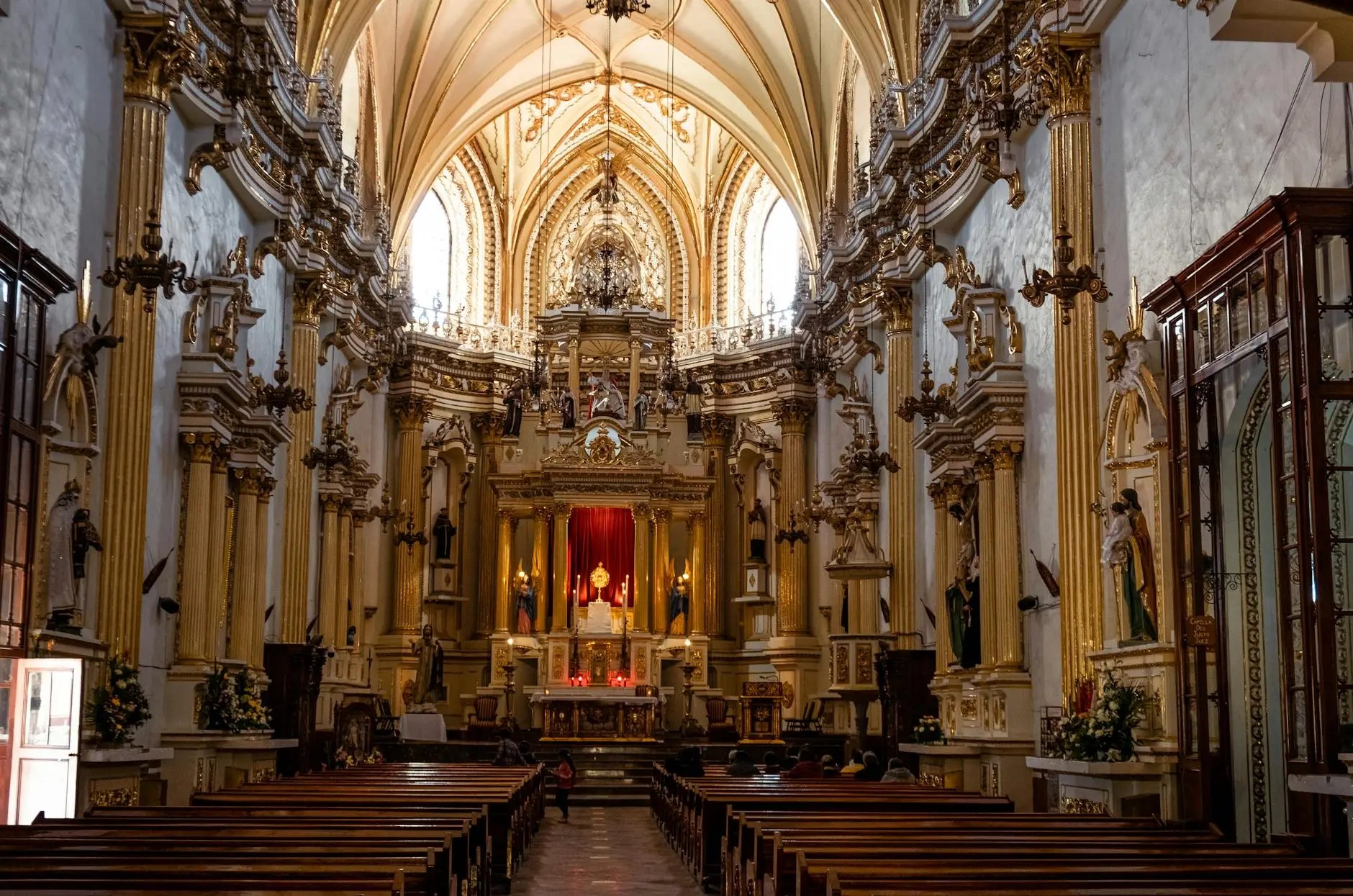 Interior of a grand church featuring ornate gold decorations, intricate architecture, and a vibrant altar, with pews in the foreground.