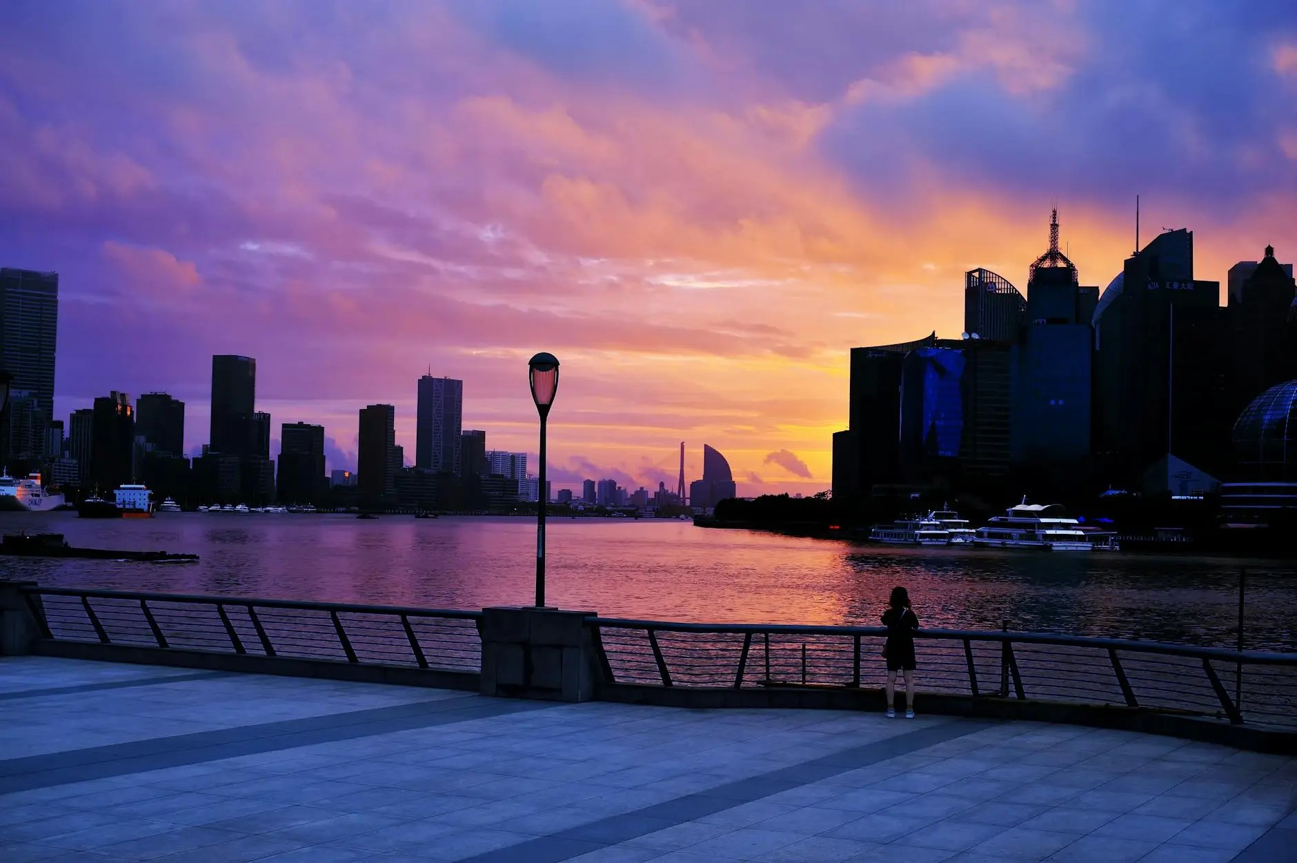 A scenic view of the Shanghai skyline at sunset, with vibrant purple and orange hues in the sky and a person standing by the waterfront.