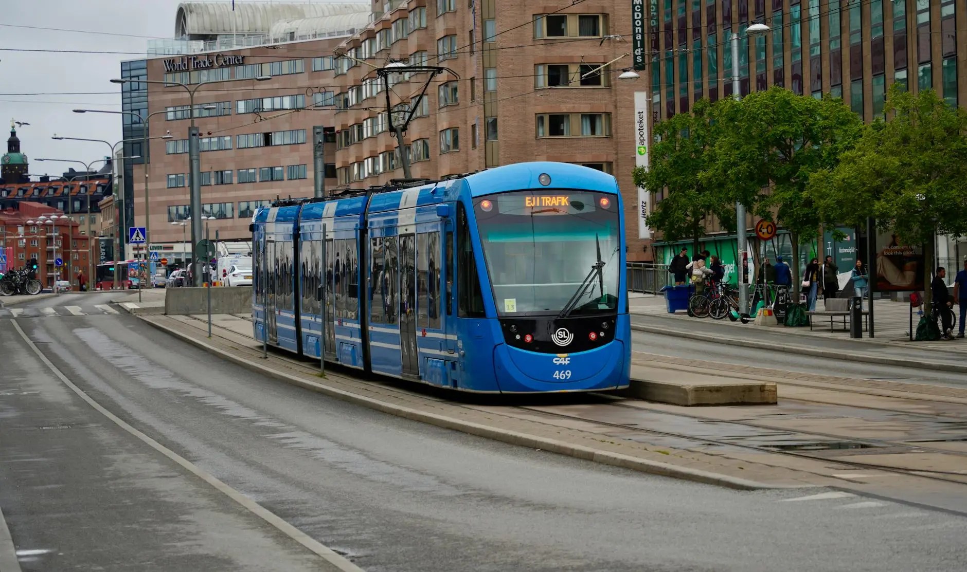 A blue tram marked 'EJ I TRAFIK' on the tracks in a modern urban setting, with buildings and trees in the background.