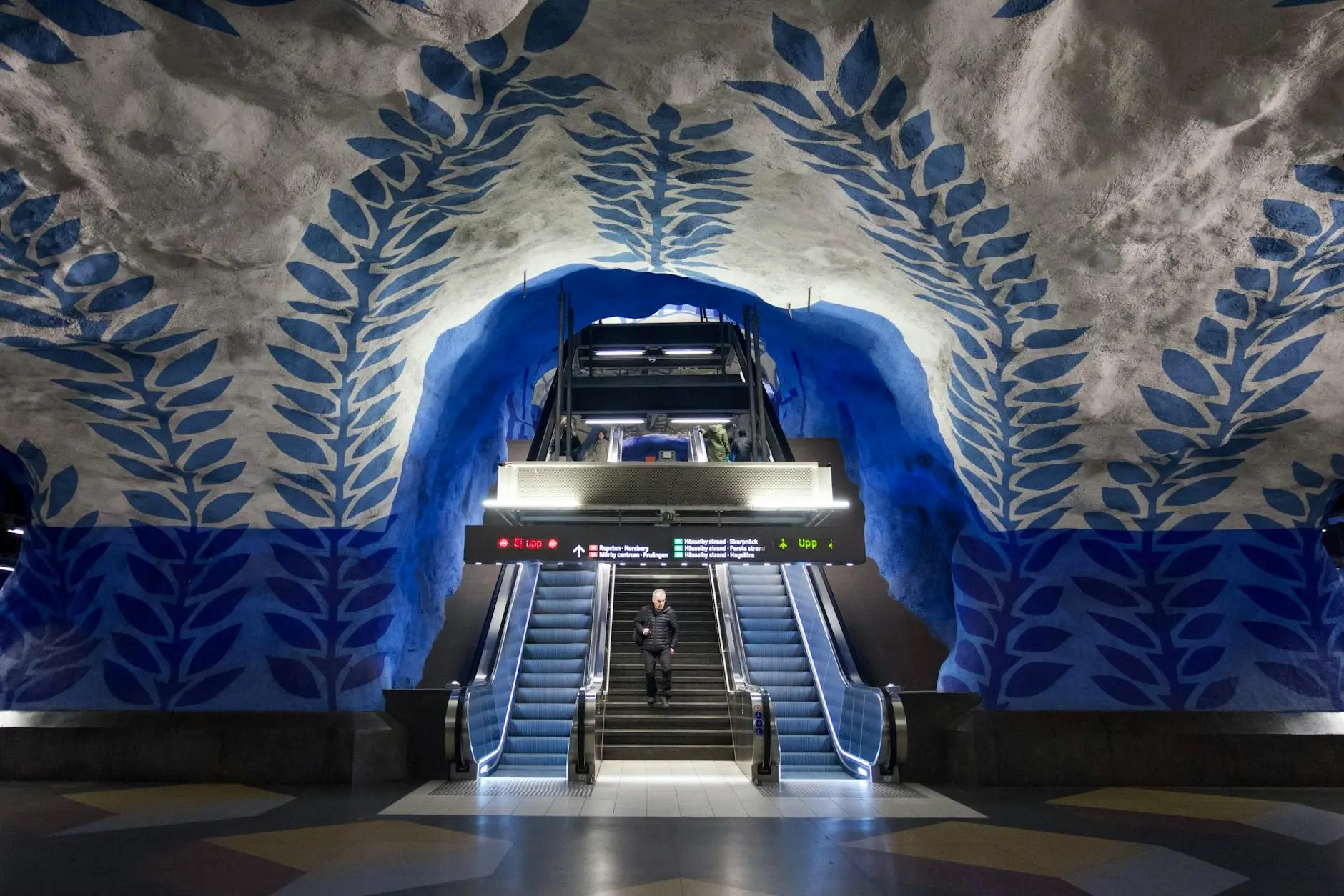 Interior of a Stockholm subway station featuring colorful blue and white mural designs on the rock walls, with stairs and escalators leading to the platform.