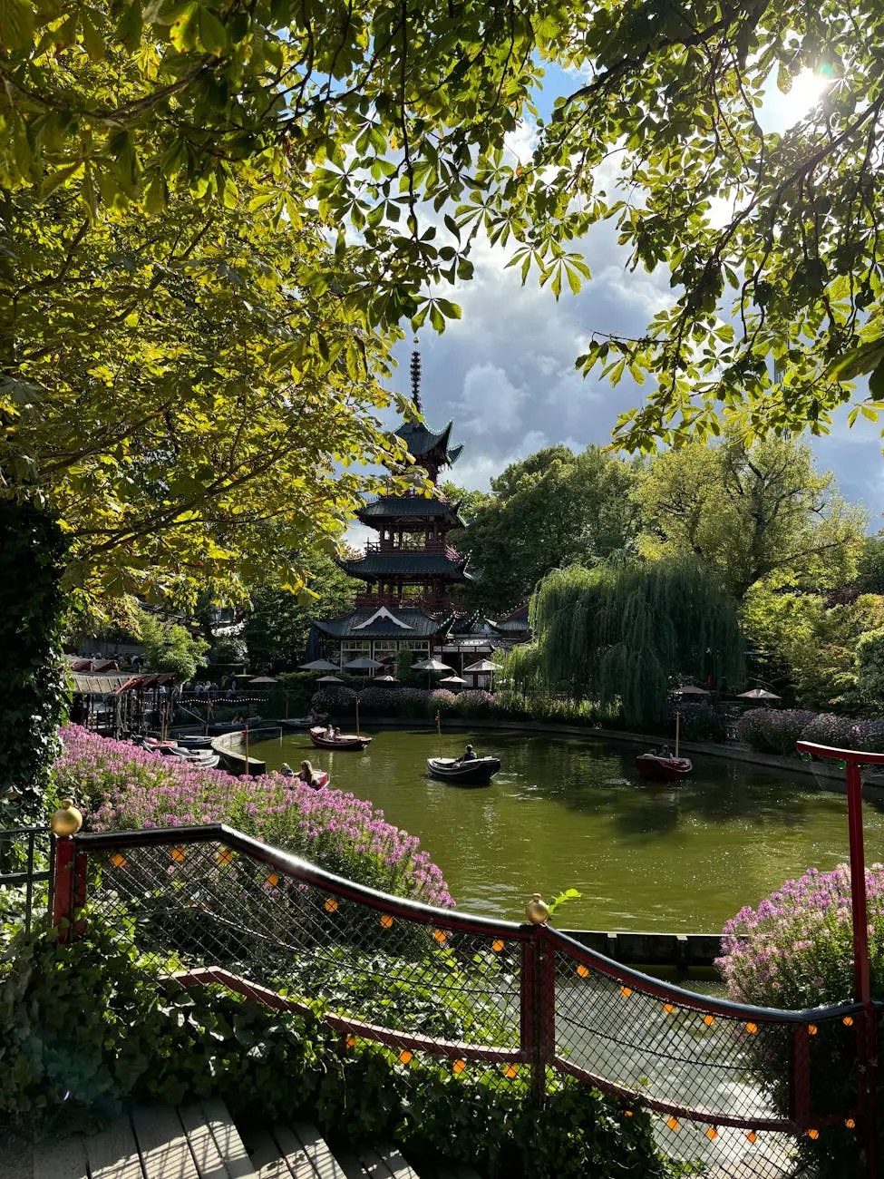A serene view of a picturesque garden with lush green trees, colorful flowers, and a tranquil pond, featuring a traditional pagoda-style structure in the background.