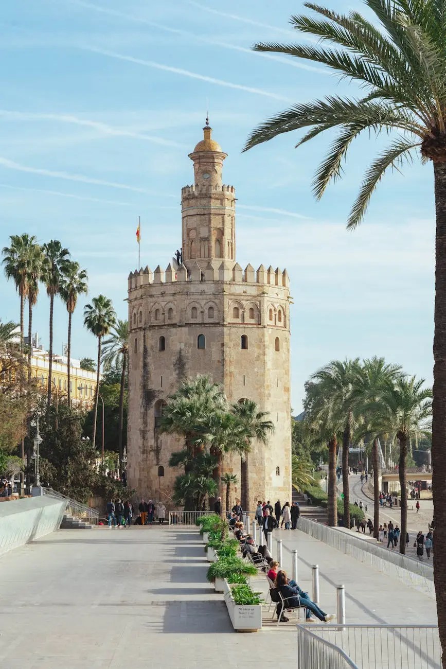 A historic tower with a golden dome, surrounded by palm trees and people walking along a promenade in Seville, Spain.
