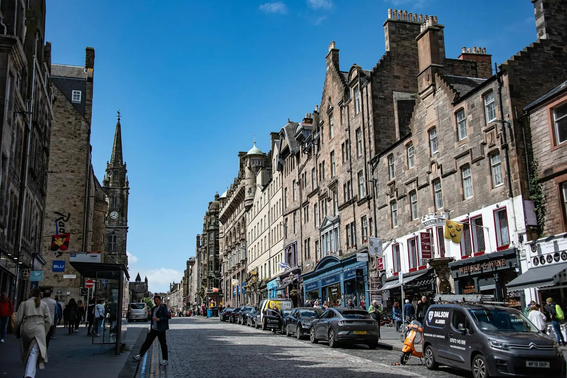 A busy street scene in Edinburgh, Scotland, featuring historic buildings, cars parked along the cobblestone road, and people walking. The sky is clear and blue, providing a vibrant backdrop to the architectural detail.