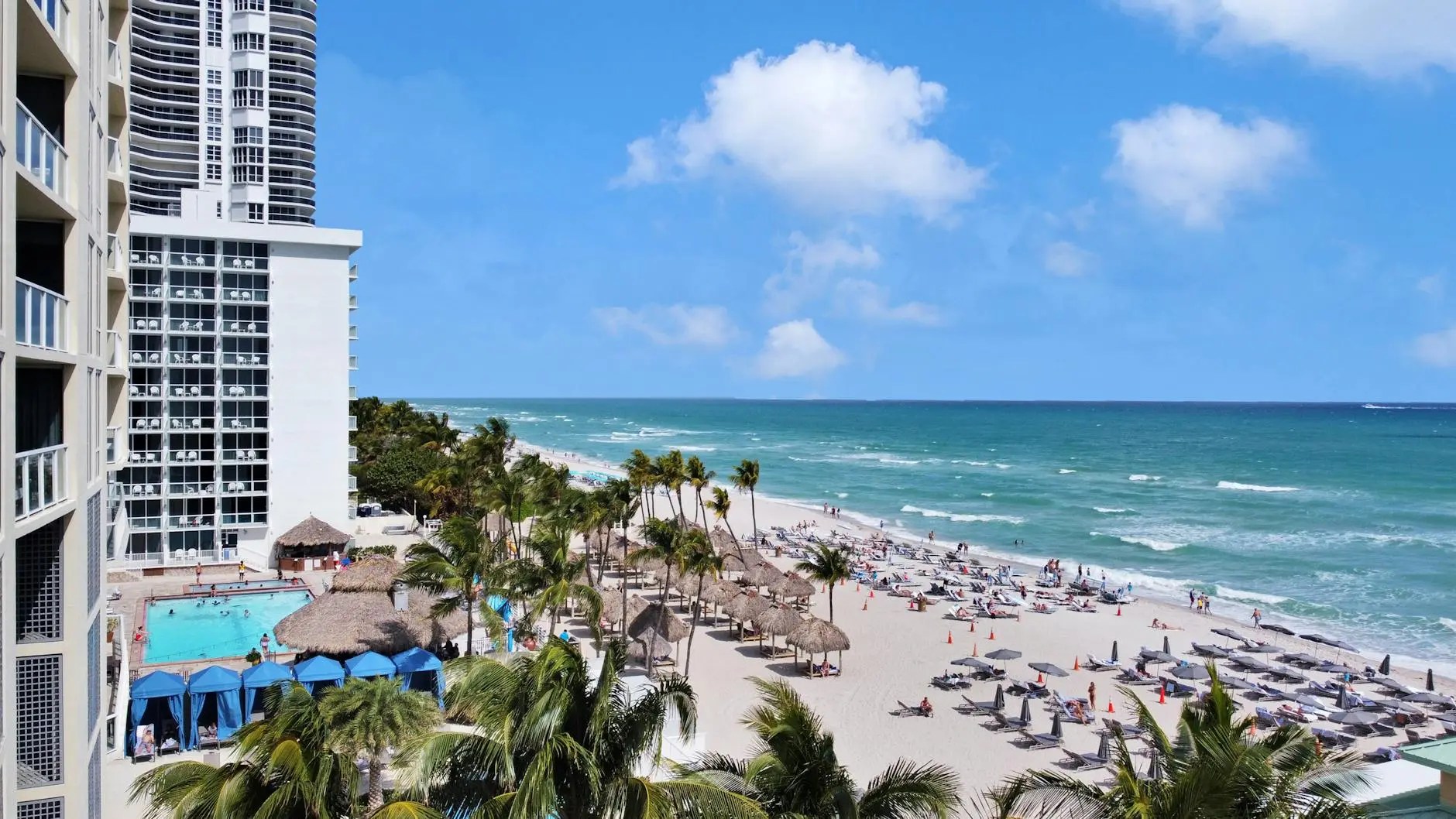 A view of a sunny Miami beach featuring turquoise waters, palm trees, and beach umbrellas, with a hotel in the foreground.