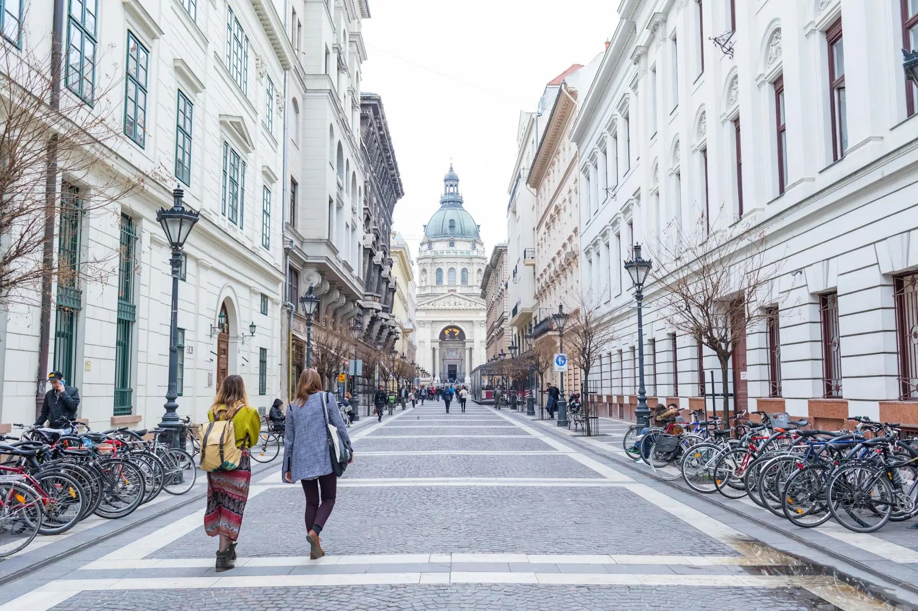 Street view in Vienna showing pedestrians walking along a cobblestone path lined with trees and bicycles parked on the side, with historic buildings and a dome structure in the background.