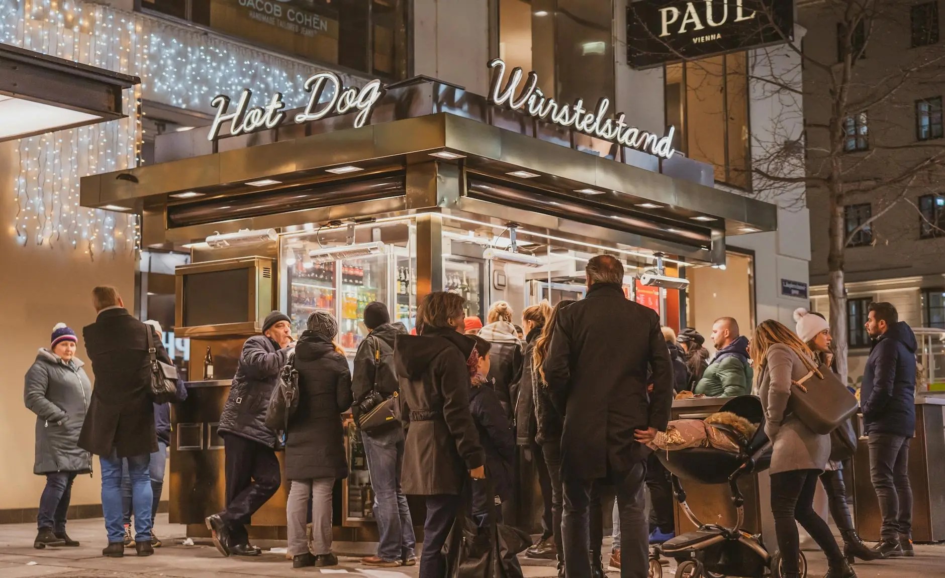 A bustling Würstelstand (sausage stand) in Vienna at night, with customers waiting in line for traditional snacks, illuminated by warm lights.
