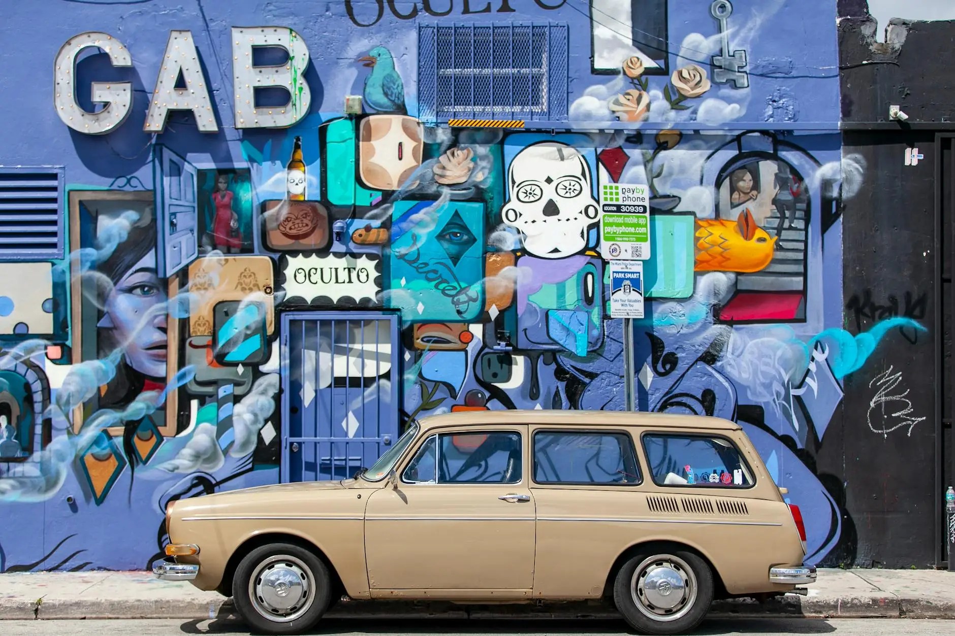 A beige vintage car parked in front of a colorful mural with various artistic elements including faces, symbols, and a door, set against a bright blue background.