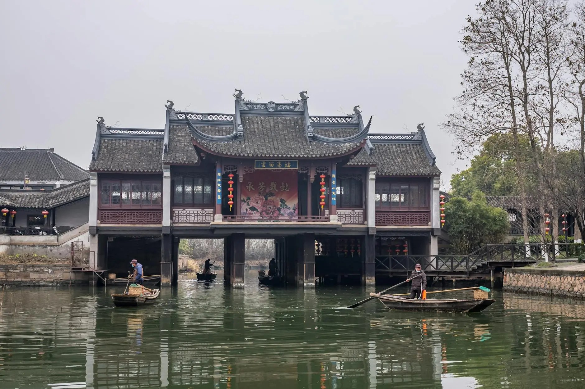 A traditional wooden building with a curved roof sits over a serene waterway, where two people are rowing boats beneath it. The scene reflects a peaceful water town atmosphere in Shanghai.