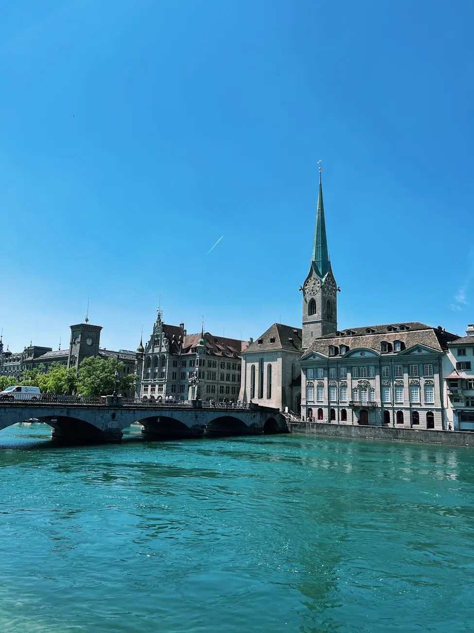 A picturesque view of Zurich featuring the turquoise waters of the Limmat River, a historic bridge, and iconic buildings with a tall church steeple under a clear blue sky.