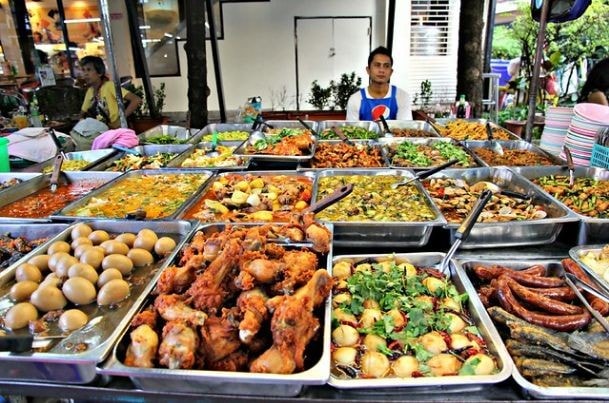 A vibrant street food market in Manila, featuring various dishes displayed in stainless steel trays, with a vendor seated in the background.