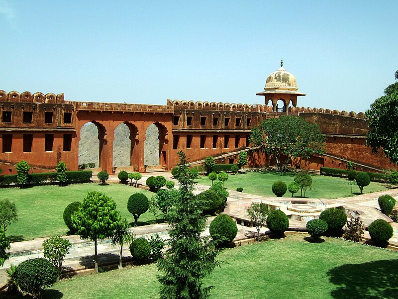 View of a landscaped garden with neatly trimmed bushes and a historic fort structure in the background, under a clear blue sky.