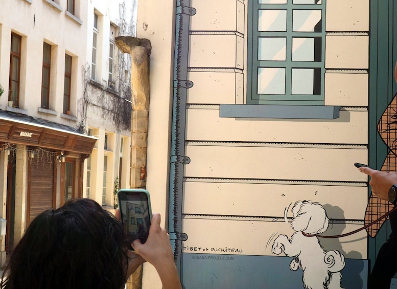 A person photographing a comic mural depicting a small dog on a wall, surrounded by typical Brussels architecture.