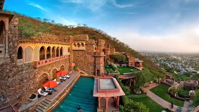 A panoramic view of a historic stone palace in Jaipur, India, featuring a swimming pool and lush greenery in the foreground.