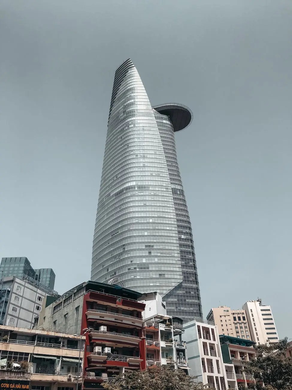 A modern skyscraper in Ho Chi Minh City, towering over older low-rise buildings, showcasing a blend of contemporary architecture against a gray sky.