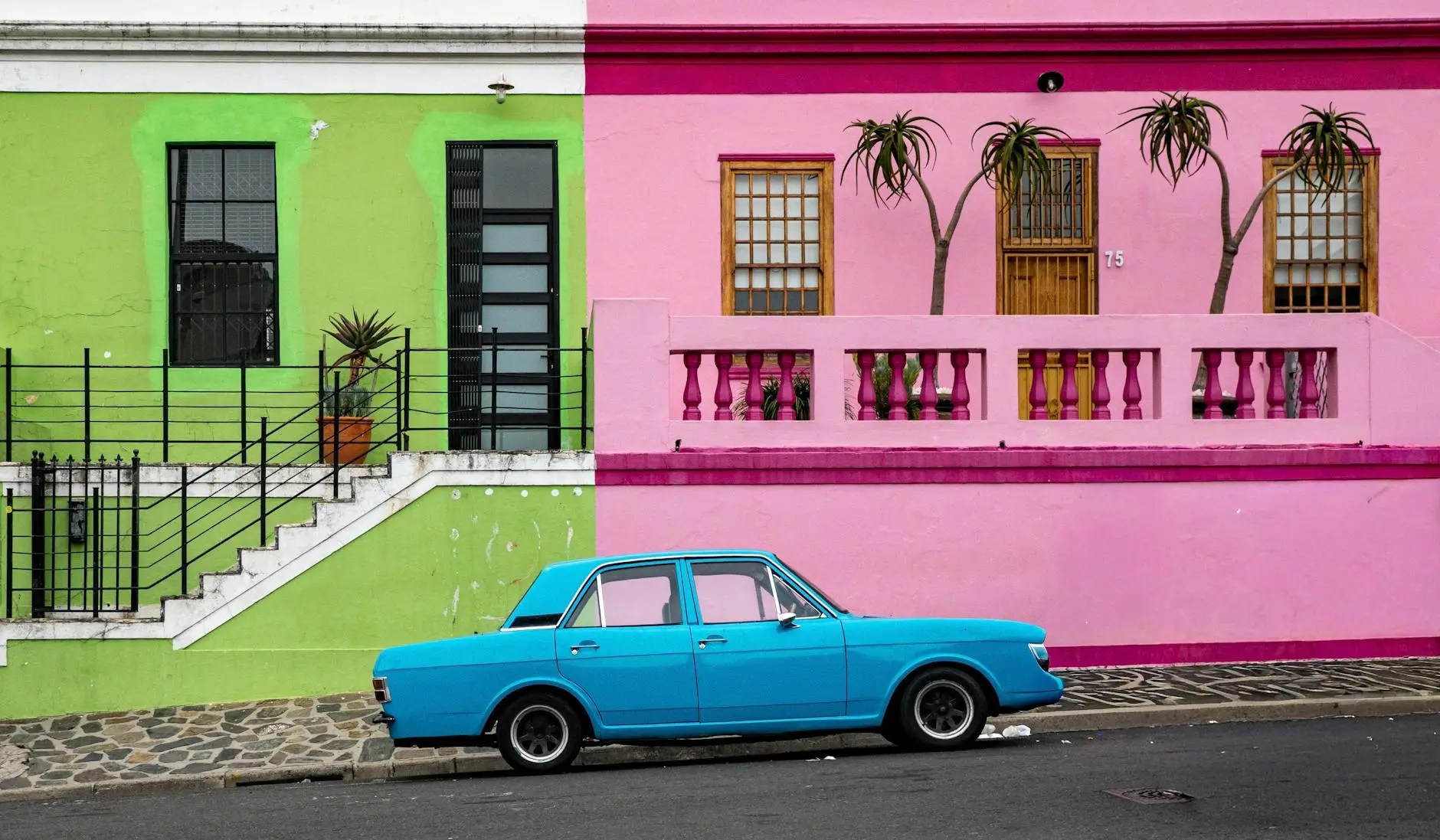 A vibrant street scene featuring a turquoise car parked beside a green and pink building, showcasing colorful architecture typical of Cape Town.