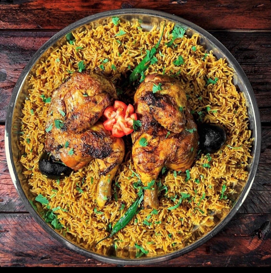 A traditional dish featuring two pieces of spiced grilled chicken served on a bed of flavorful yellow rice, garnished with fresh herbs and slices of tomato.