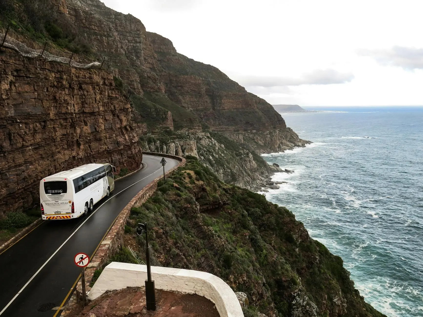 A scenic coastal road view with a bus traveling along the winding route, surrounded by steep cliffs and the ocean in the background.